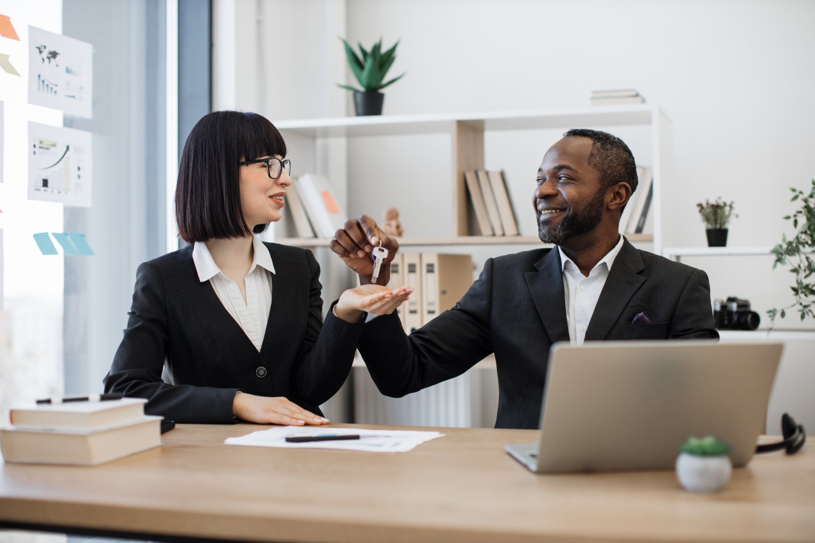 Joyful multiracial businessman in formal attire giving house keys to his caucasian female coworker while sitting behind writing desk in office. Real estate professionals finding buyers perfect home.